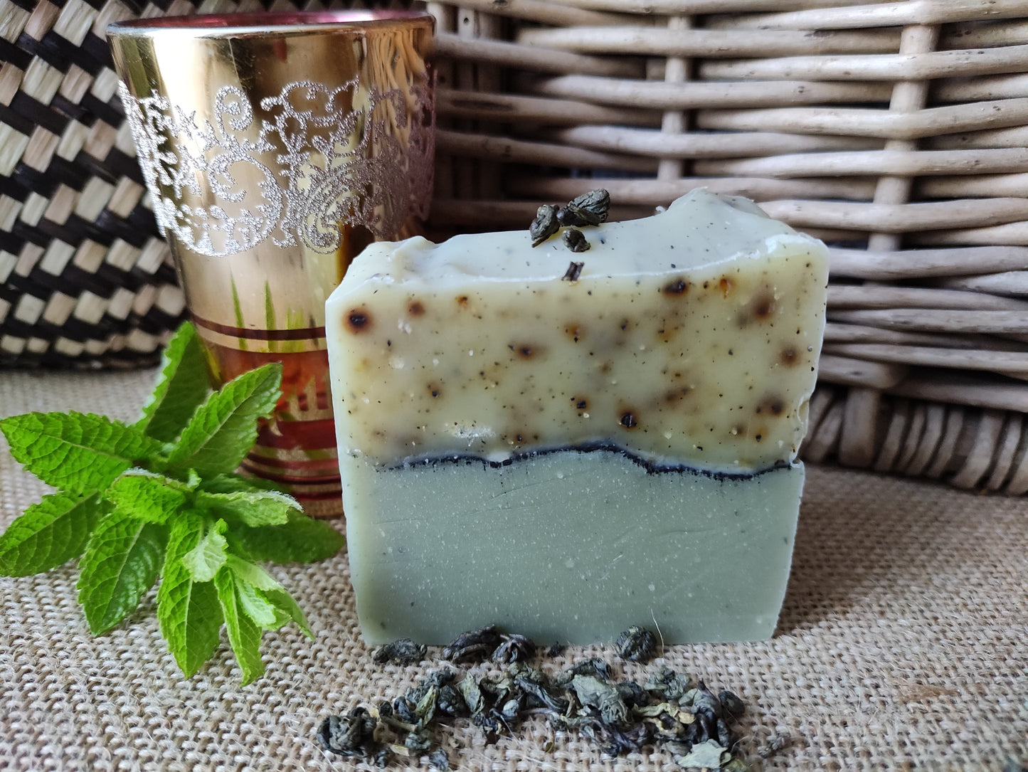 Savon artisanal saponifié à froid. Il est bicolore. La partie inférieure est vert clair et la partie supérieure blanche mouchetée de petits fragments de thé vert. une ligne de charbon sépare les deux parties. Le savon est présenté près d'un vert de thé à la menthe marocain et de feuilles de menthe fraiches
