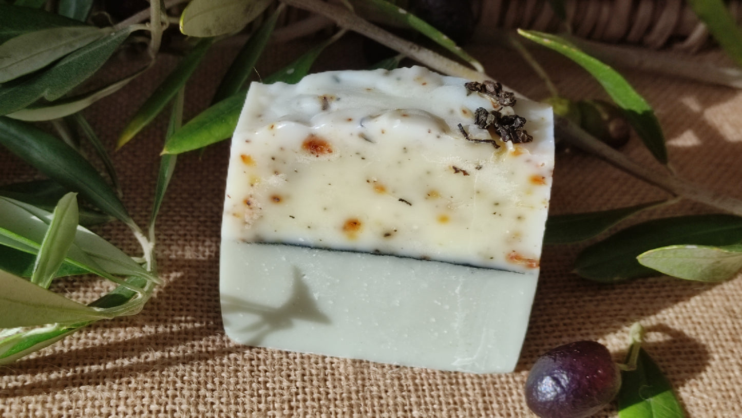 Savon artisanal saponifié à froid. Il est bicolore. La partie inférieure est vert clair et la partie supérieure blanche mouchetée de petits fragments de thé vert. une ligne de charbon sépare les deux parties. Le savon est présenté à côté d'une branche d'olivier portant des olives