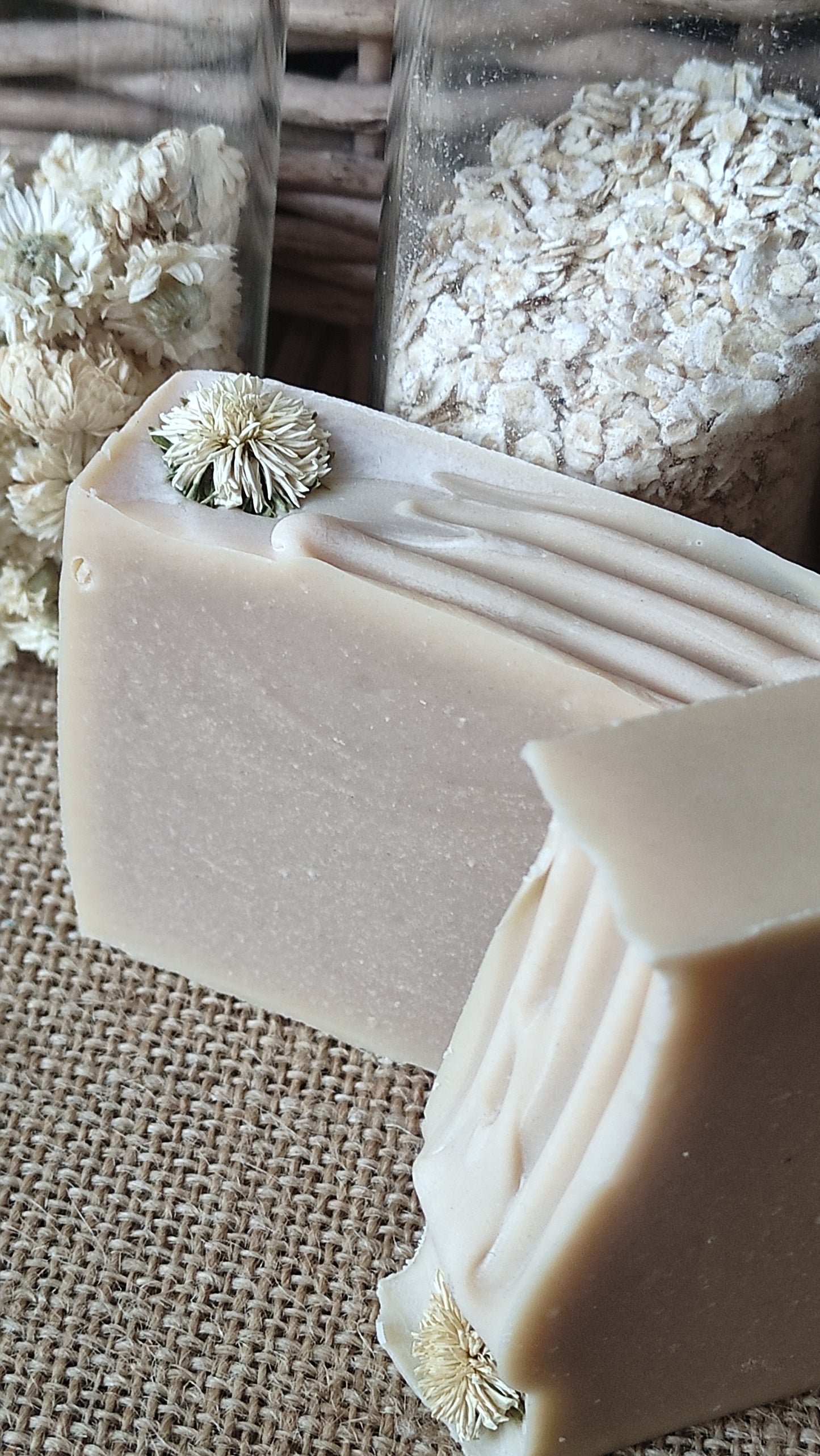 Savon artisanal saponifié à froid. Il est rose pâle avec ne fleur de camomille sur le dessus. Flacons d'avoine et de fleurs de camomille en arrière plan