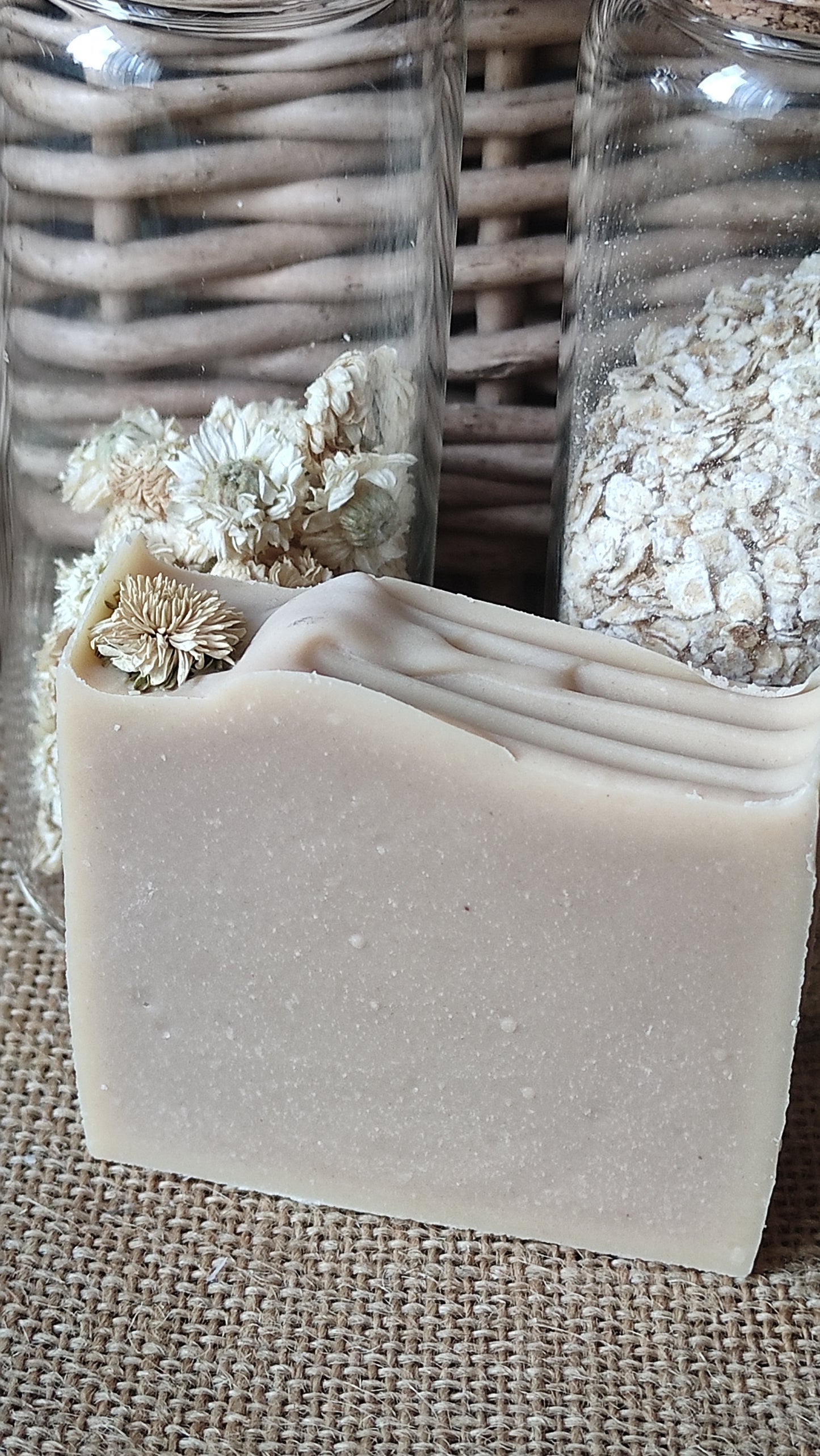 Savon artisanal saponifié à froid. Il est rose pâle avec ne fleur de camomille sur le dessus. Flacons d'avoine et de fleurs de camomille en arrière plan