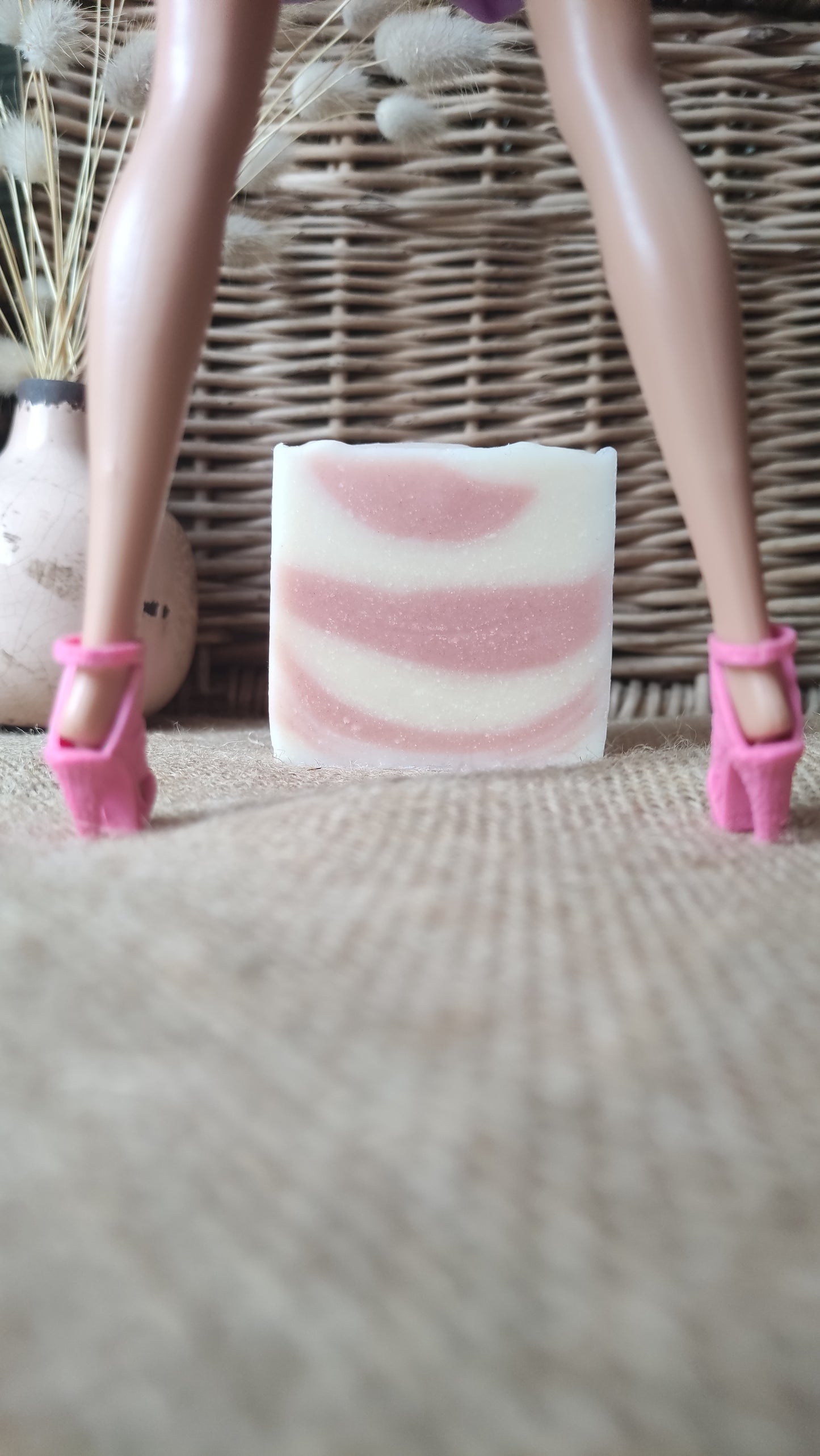 savon surgras saponifié à froid au lait d’ânesse. Il est strié rose et blanc et est présenté en arrière plan entre les jambes d'une poupée Barbie