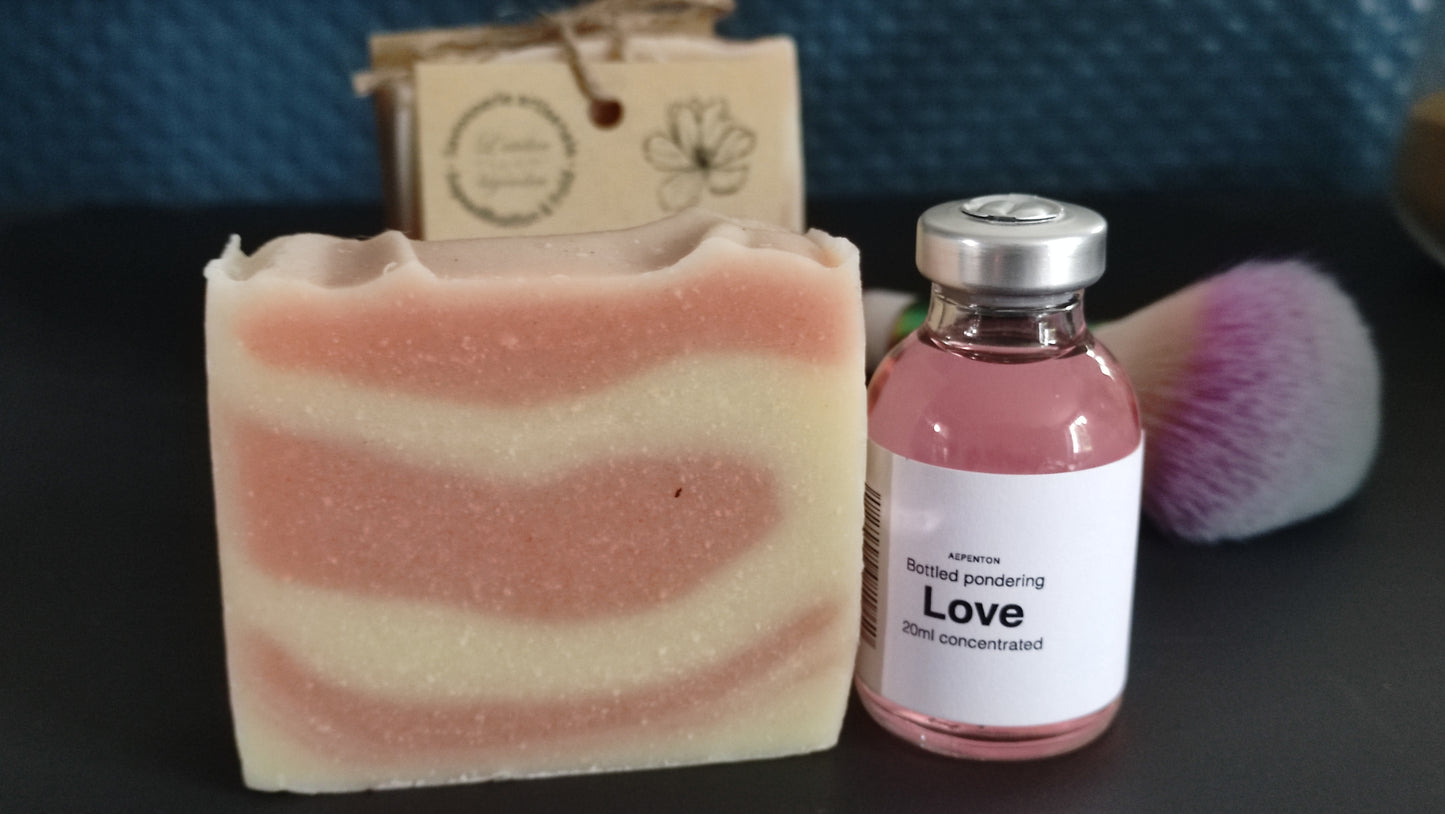 savon surgras saponifié à froid au lait d’ânesse. Il est strié rose et blanc et est présenté à côté d'une fiole Love de Aepenton