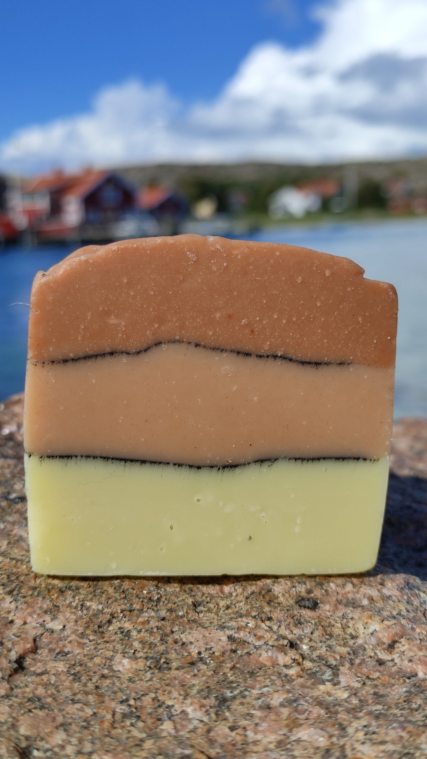 savon surgras saponifié à froid à l'huile de pépin de raisin. Il est composé de trois couches séparée par ne ligne de charbon. La première est couleur beige, la deuxième rose pâle et la dernière rose plus foncé. Le savon est posé sur un rocher devant des maisons rouges suédoises