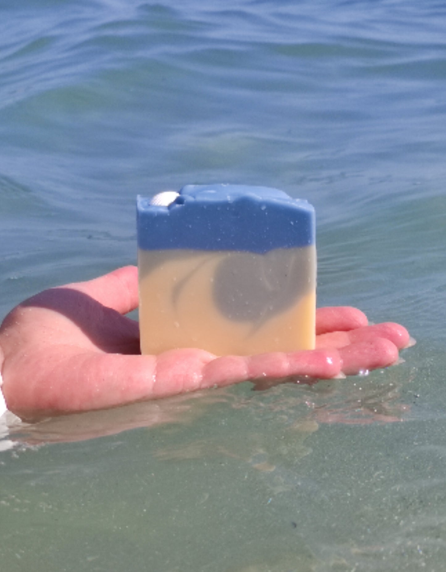 Savon saponifié à froid avec des dégradés bleus et blancs rappelant la mer. Un coquillage blanc est posé dessus. Le savon est dans un main au ras de l'eau de mer
