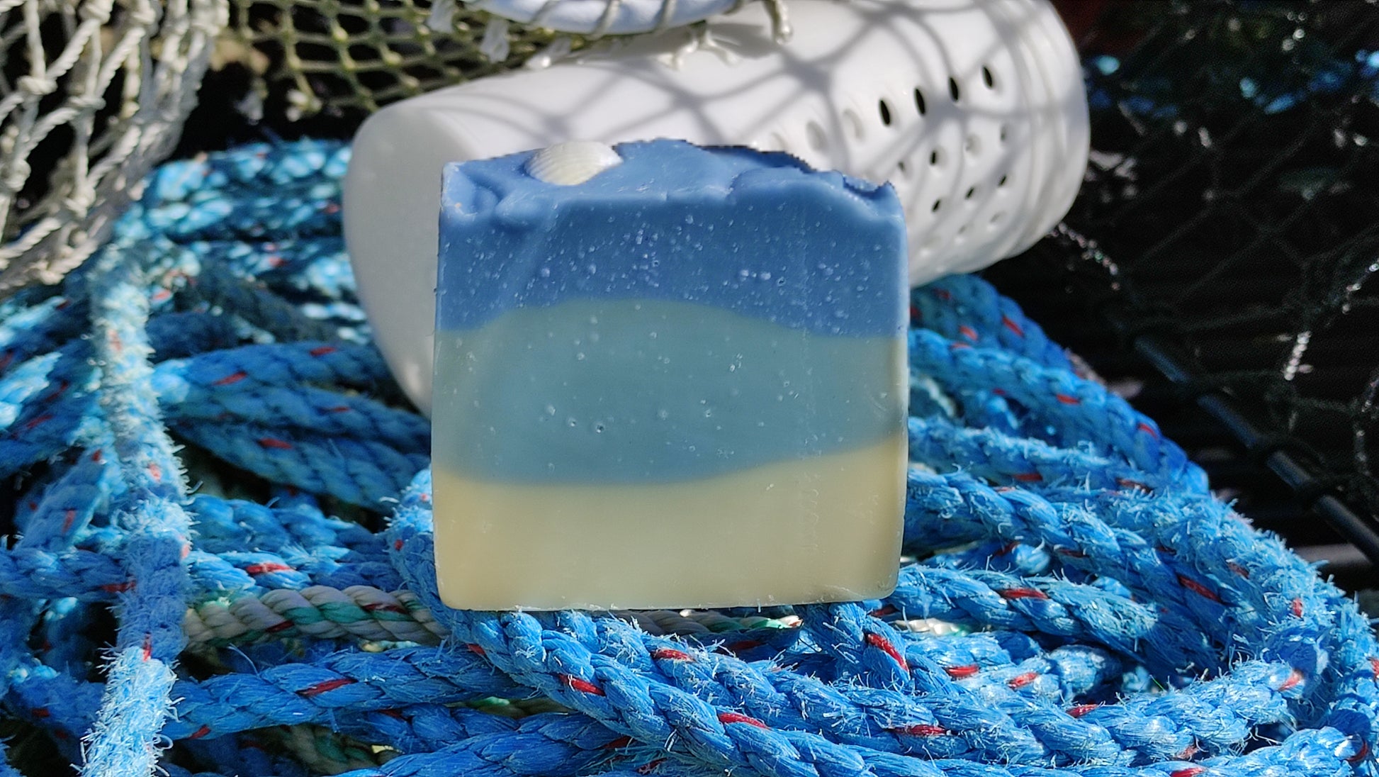 Savon saponifié à froid avec des dégradés bleus et blancs rappelant la mer. Un coquillage blanc est posé dessus. Le savon est au milieu de filets de pêche.