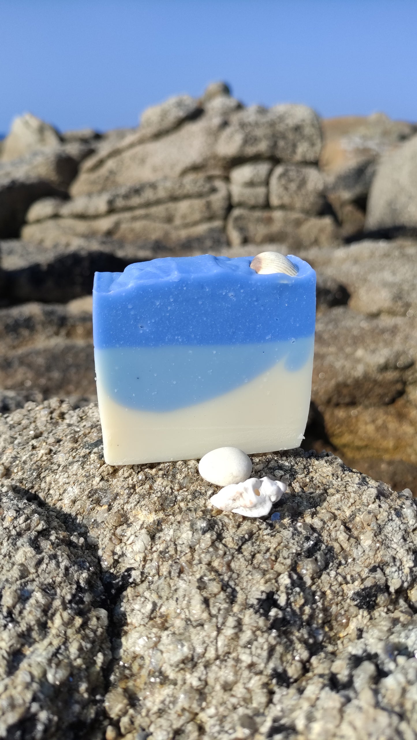 Savon saponifié à froid avec des dégradés bleus et blancs rappelant la mer. Un coquillage blanc est posé dessus. Le savon est posé sur un rocher au bord de la mer
