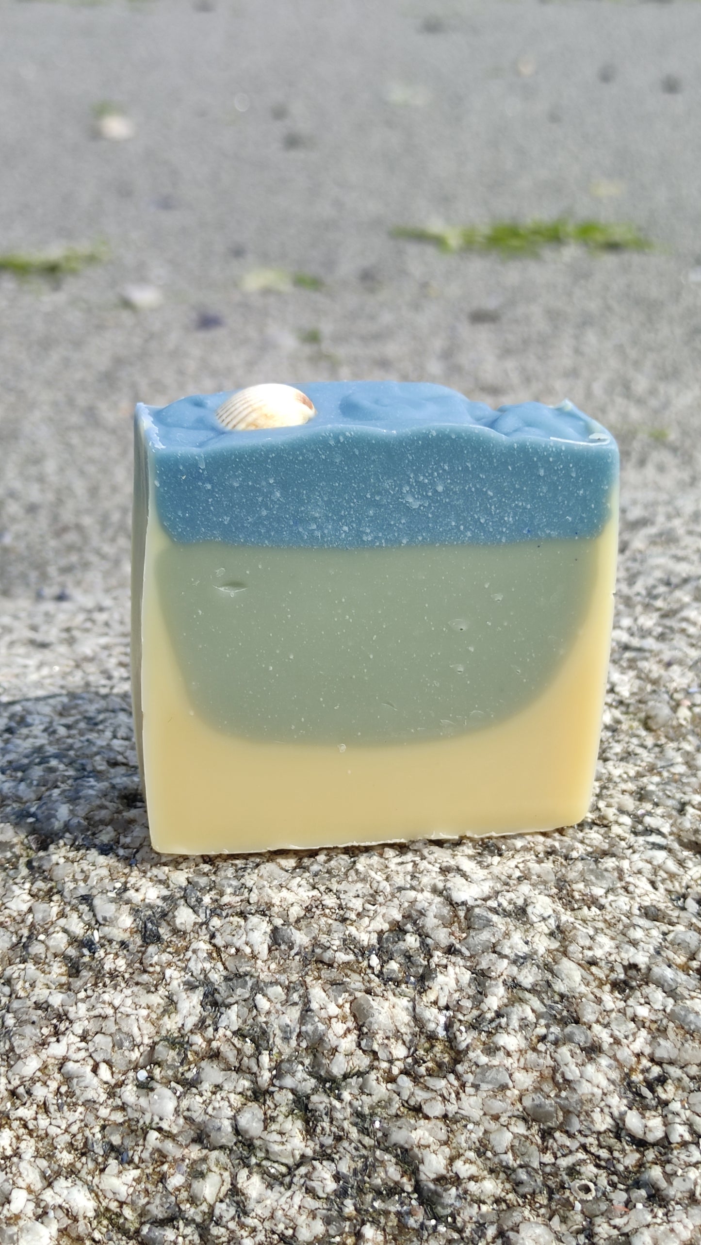Savon saponifié à froid avec des dégradés bleus et blancs rappelant la mer. Un coquillage blanc est posé dessus. Le savon est sur la plage.