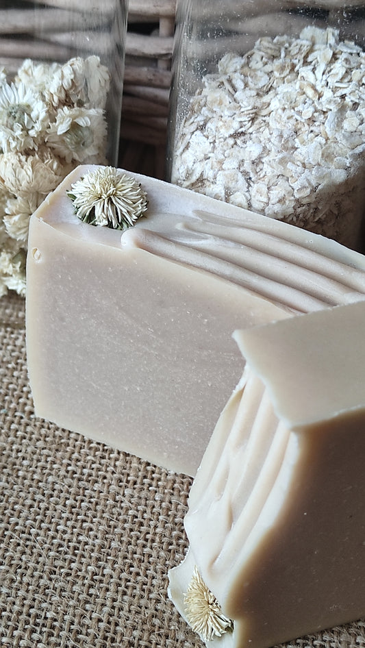 Savon artisanal saponifié à froid. Il est rose pâle avec ne fleur de camomille sur le dessus. Flacons d'avoine et de fleurs de camomille en arrière plan
