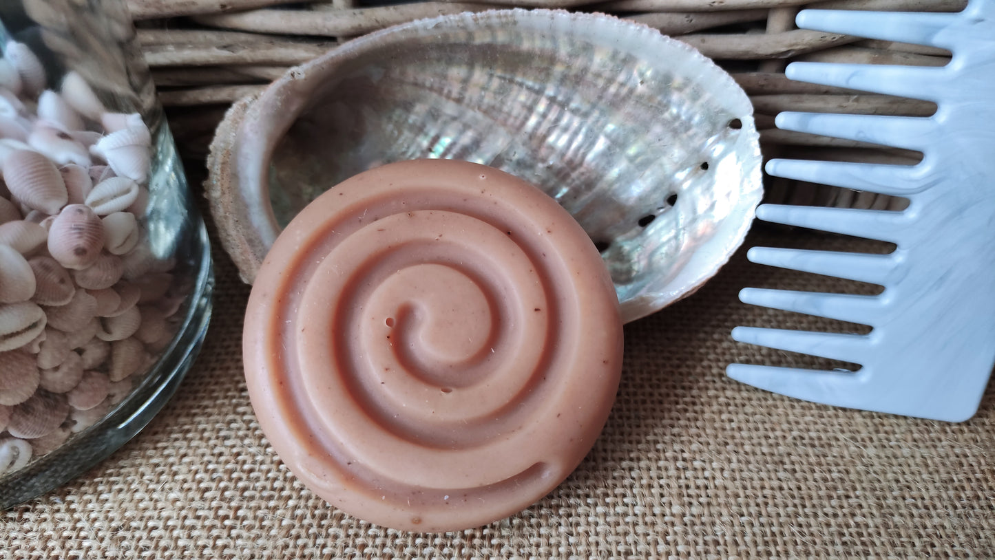 shampooing naturel de forme ronde avec une spirale dessus posé sur une coquille d'ormeau avec peigne en filet de pêche recyclé à côté