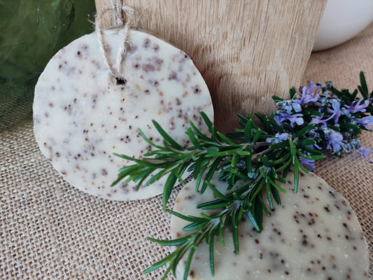 savon disque rond traversé par une ficelle de chanvre pour le suspendre. Il est de couleur crème et moucheté de marron (grains de fèves de café exfoliante) et présenté avec une branche de romarin